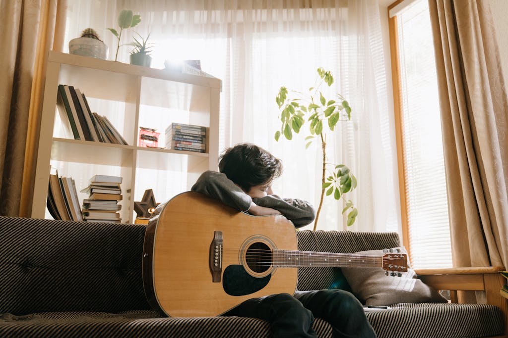 A young boy rests with his acoustic guitar at home, surrounded by books and plants, embracing a peaceful moment.