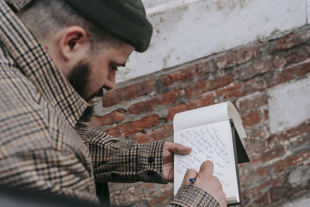 A man in a plaid coat writes lyrics in a notebook by a brick wall outdoors.