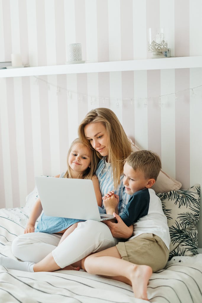 A mother sits on a bed with her children, sharing a joyful moment using a laptop.