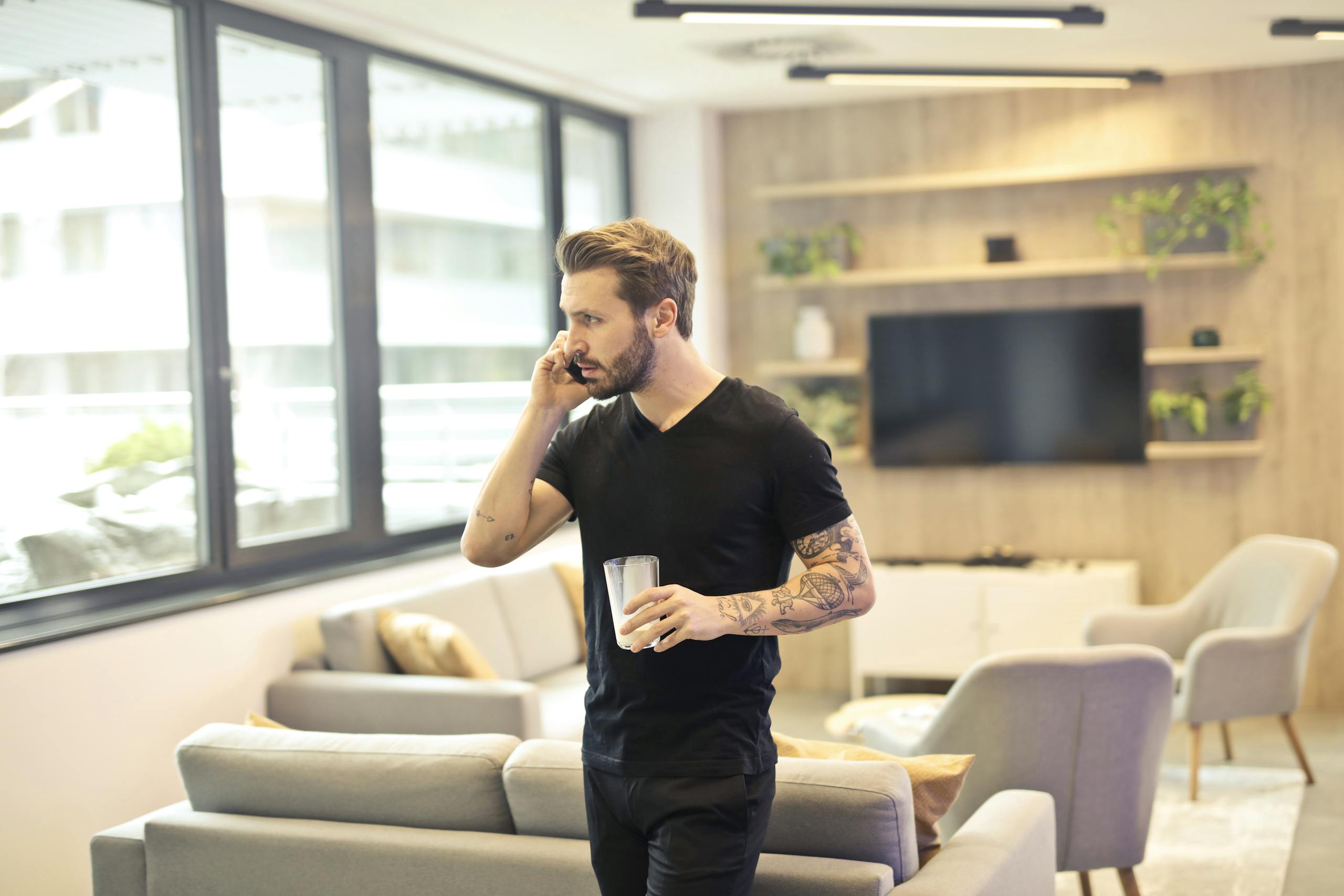 A tattooed man talking on a smartphone and holding a drink in a contemporary living room setting.