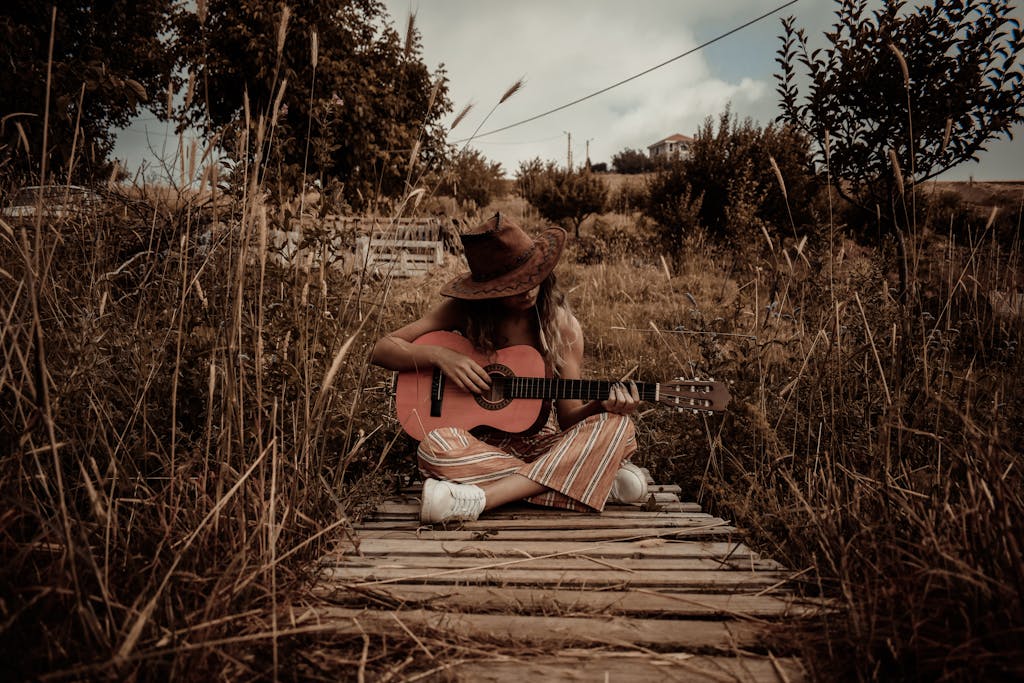 A woman in a hat playing an acoustic guitar in a rural, grassy landscape, exuding a peaceful, rustic vibe.