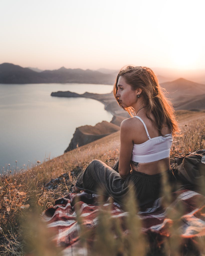 A woman sits on a hillside overlooking a tranquil lake at sunset, surrounded by nature.