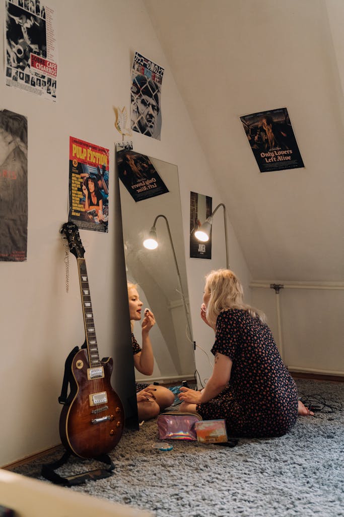 A young Caucasian woman applies makeup in a cozy room with posters and a guitar.