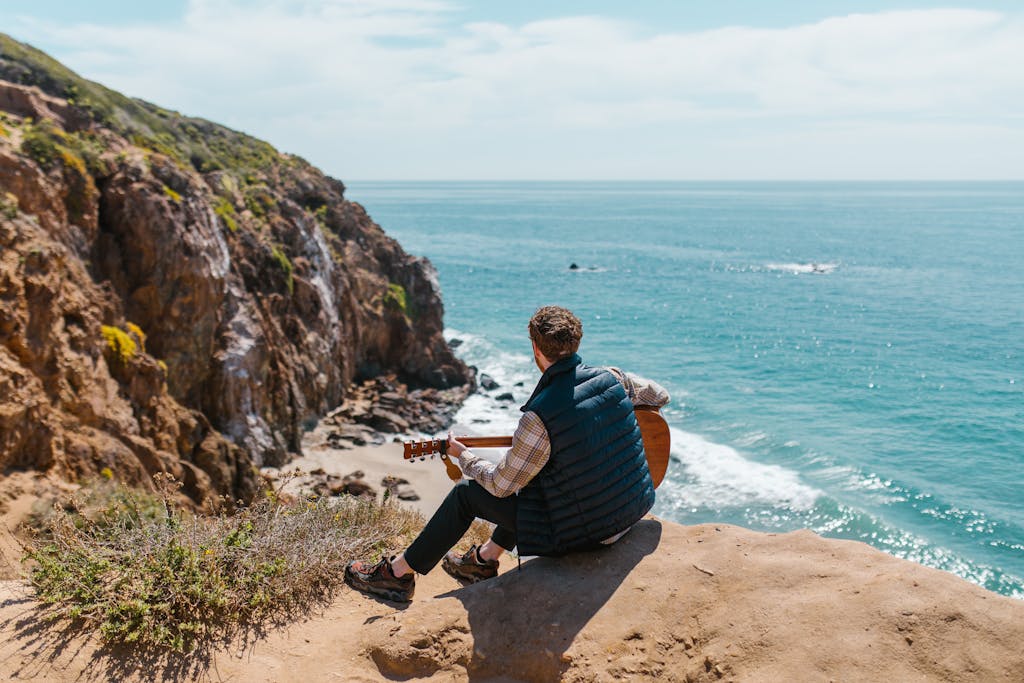 An adult with a guitar sits on a cliff overlooking the ocean, embracing nature and serenity.