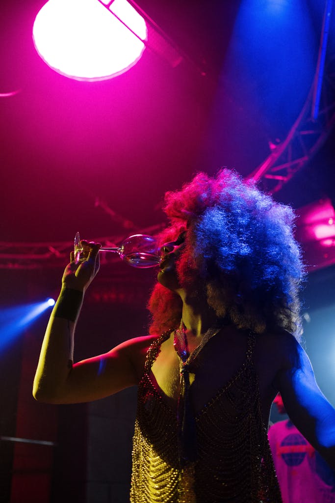 Colorful performer with afro hairstyle drinks from a glass on a vibrant nightclub stage.
