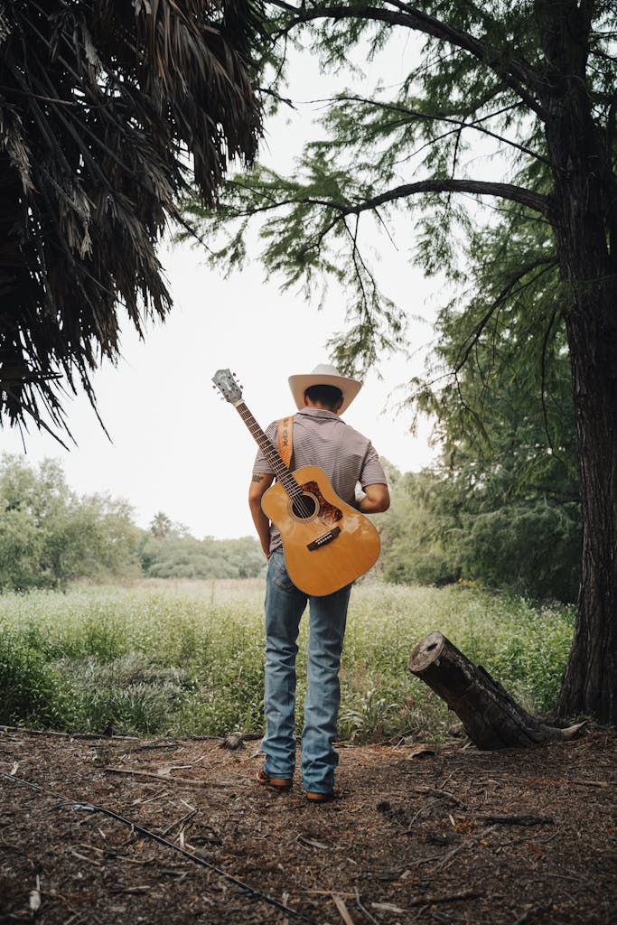 Man in cowboy hat with guitar standing in serene forest clearing, captured from behind.