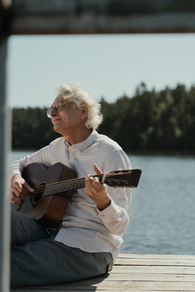 Senior man playing acoustic guitar on a sunny lakeside pier, enjoying a peaceful moment.