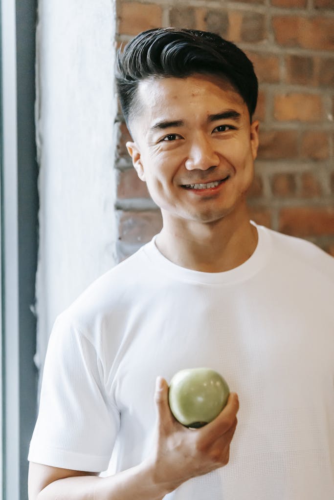 Smiling man indoors holding a green apple, promoting healthy eating and positivity.