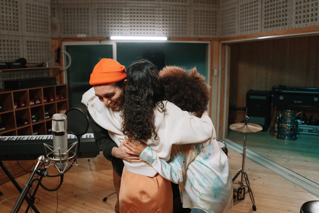 Three people warmly embrace in a music recording studio, showcasing friendship and teamwork made better by therapy with Musician Therapy Collective