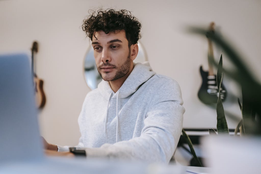 Young unshaven male musician in hoodie surfing internet on netbook against musical instruments at home